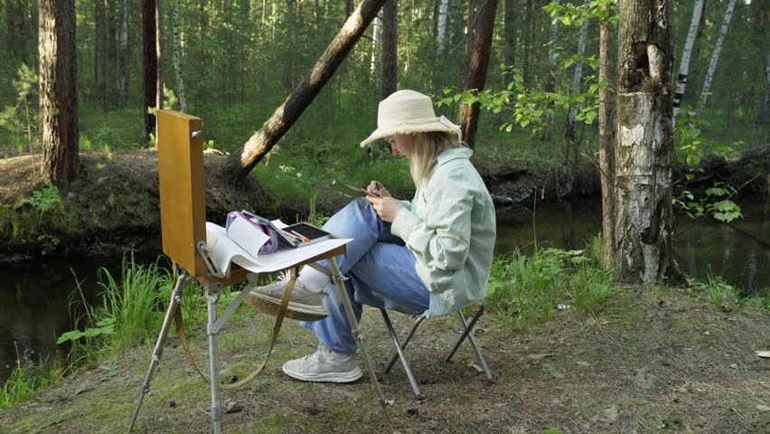 A romantic girl artist, inspired by the forest nature, paints a landscape by a forest river. Mosquitoes bite and do not give rest. The creative process.