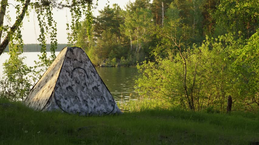 A tourist tent on the shore of a lake in a blooming green forest. Hiking, camping, fishing, healthy lifestyle. Birds are flying on the lake.