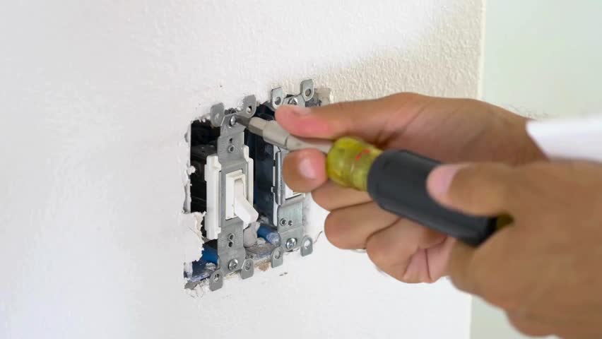 electrician fixing a wall-mounted light switch, ready to toggle it. The room is illuminated with natural light.