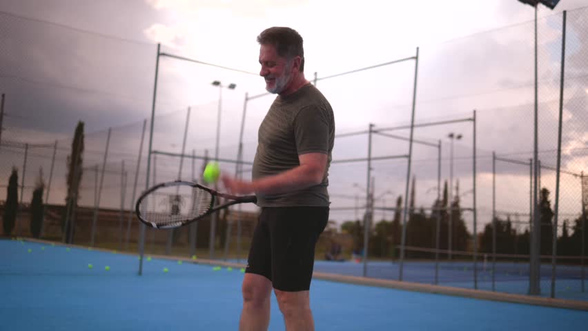 Mature male tennis player practicing powerful serve on blue tennis court, demonstrating athletic skill and concentration during evening hours under dusky sunset sky