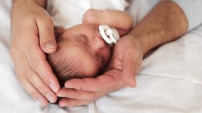 Close-up video shows a father's hands tenderly cradling his sleeping newborn baby with a pacifier on a white sheet. The scene emphasizes paternal love and gentle care - Powered by Shutterstock - Get 15% off with code: PIKWIZARD15