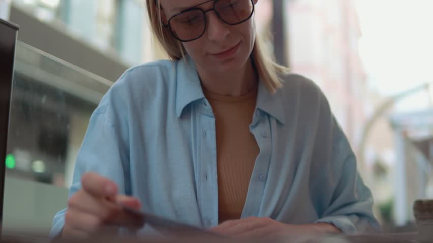 Young woman reading restaurant menu, wearing blue shirt and glasses, carefully selecting dining options with contemplative look at table