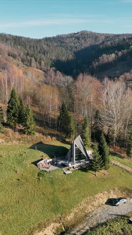 Hillside A-Frame Cabin with Scenic Views. Aerial view of an A-frame cabin on a grassy hillside, surrounded by trees and rolling hills under a clear blue sky.