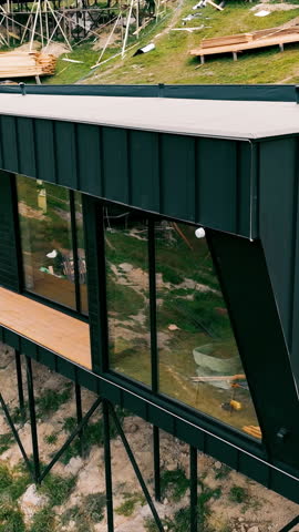 Elevated modern chalet on stilts with hillside views. A modern black cabin with large glass windows, elevated on tall metal stilts above a grassy hillside, featuring a wooden balcony under