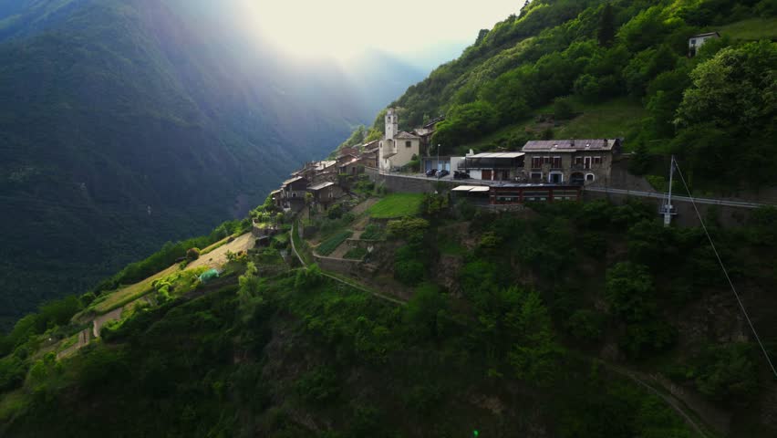 Drone shot of a cliffside village road winding past alpine homes and terraced fields with dramatic mountain drop-offs. Shot at Tirano, Italy (Tirano, Italia)