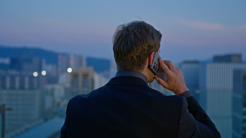Caucasian businessman in a suit turns while talking on phone, overlooking a city skyline at sunset from a rooftop terrace, - dolly back revealing shot