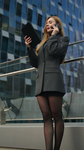 Professional businesswoman stands on a terrace, holding a tablet and talking seriously on her phone, surrounded by modern glass office buildings - parallax shot