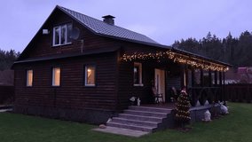 In the evening twilight on Christmas Eve, garlands are lit on the terrace of the wooden cottage and on the thuja growing by the porch. A cat is walking. A winter without snow and green lawn grass - Powered by Shutterstock - Get 15% off with code: PIKWIZARD15