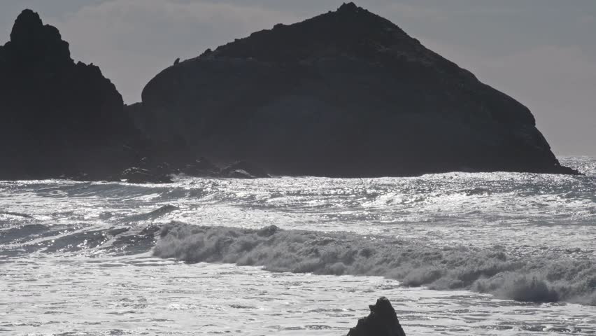 Powerful waves crash against a large rock formation silhouetted by the evening sun on a rugged Pacific shoreline.