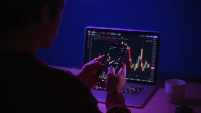 Over shoulder view of crypto trader analyzing live crypto and stock charts on smartphone with trading platform app, displayed on laptop and phone screen in dark neon lit room.