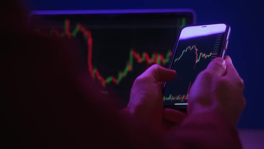 Trader hands holding smartphone, using trading app for analyzing financial, stock market and cryptocurrency data, with red and green candlestick charts on screen, in dark neon lit office.