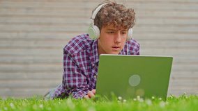 Curly teenage boy listening to music on headphones and works on a laptop while sitting in nature in a park - Powered by Shutterstock - Get 15% off with code: PIKWIZARD15