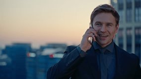 Blond Caucasian businessman in a suit smiles confidently while talking on his smartphone at an outdoor office terrace with a city skyline at sunset - Powered by Shutterstock - Get 15% off with code: PIKWIZARD15