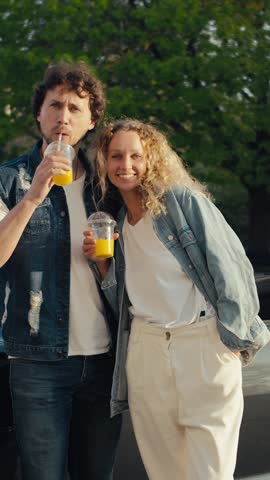 Two friends savor refreshing orange beverages while smiling in a sunny park, surrounded by green trees and a lively atmosphere, creating lasting memories