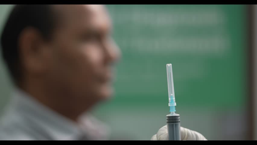 A close-up conceptual video of a nurse carefully opening the cap of a syringe, with a blurred male figure in the background. The scene emphasizes medical precision, patient care, and clinical readines