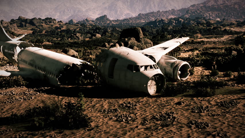 A damaged aircraft is seen partially buried in rocky ground, showing signs of a severe crash. The surrounding landscape features hills and sparse vegetation under a clear sky.