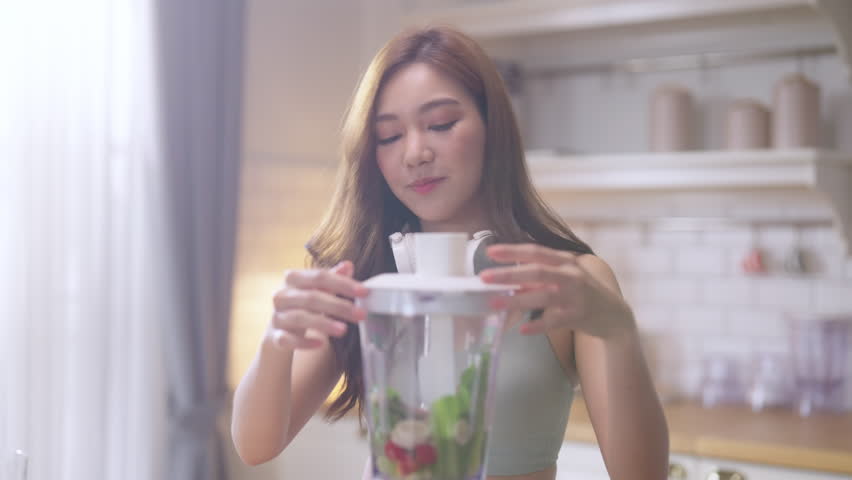 cheerful young Asian athletic woman in sporty casual wear is smiling while preparing a healthy protein shake with fresh fruits and vegetables in her modern kitchen at home, embracing a balanced diet, 