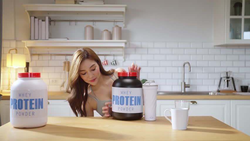 A young, athletic Asian woman in stylish sportswear prepares a nutritious protein shake in her modern kitchen while live-streaming a health-focused video tutorial. With a cheerful smile