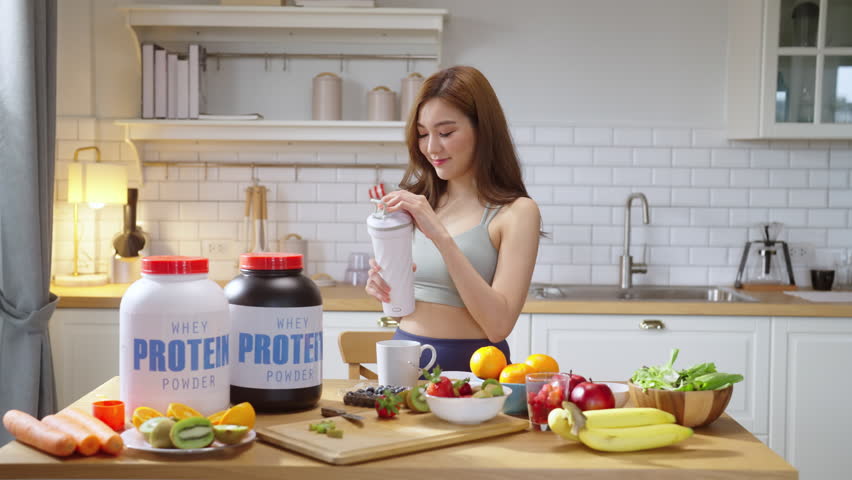 A young, athletic Asian woman in stylish sportswear prepares a nutritious protein shake in her modern kitchen while live-streaming a health-focused video tutorial. With a cheerful smile