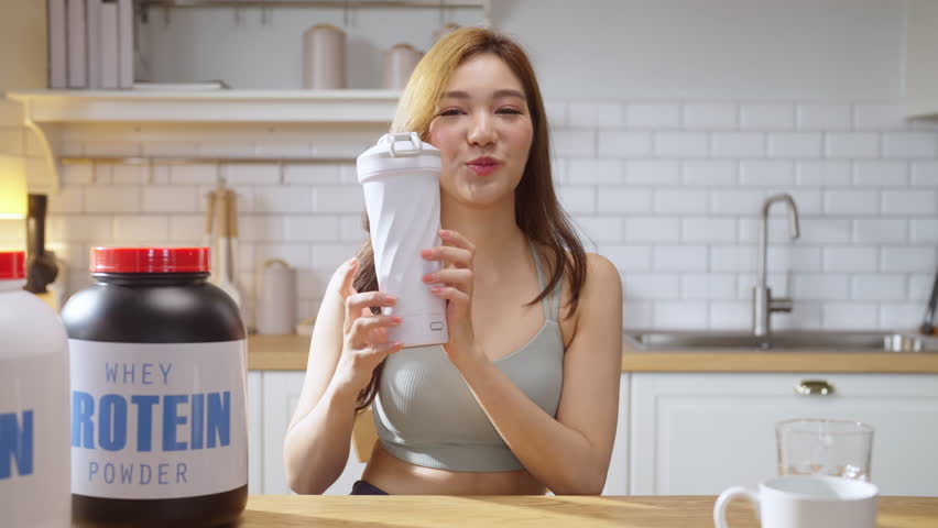 A young, athletic Asian woman in stylish sportswear prepares a nutritious protein shake in her modern kitchen while live-streaming a health-focused video tutorial. With a cheerful smile