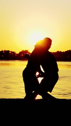 Two dark male figures, at sunrise, against the light, boxing, fighting in sparring, training in a pair of techniques of strikes, near water, in summer