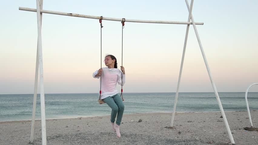 A little girl with heart-shaped glasses is swinging on a swing by the sea