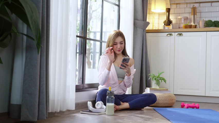 A young, healthy Asian woman in activewear takes a cheerful selfie or video call on her smartphone while relaxing after a yoga workout with her community in a cozy apartment, embracing wellness, conne