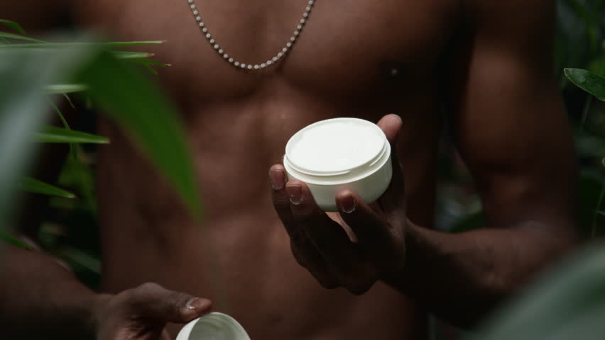 Close up shot of hands of unrecognizable shirtless man opening white cosmetic jar of skincare cream while standing in jungle