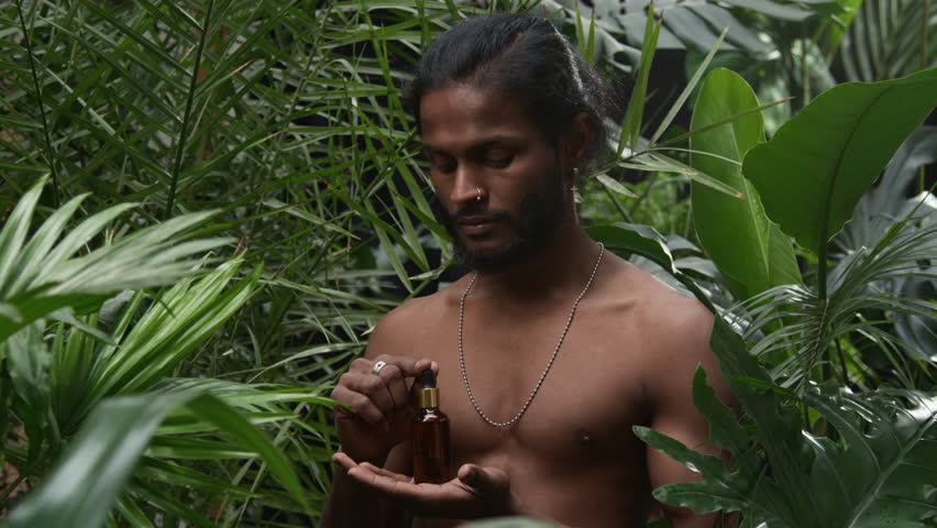 Portrait of young Indian man with bare torso standing in lush green jungle, holding dropper bottle of skin serum and looking at camera