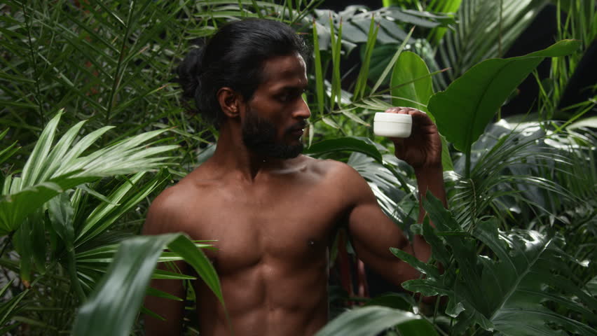 Portrait of handsome Indian man standing shirtless in lush green jungle, holding white jar of skincare cream and looking at camera with confidence