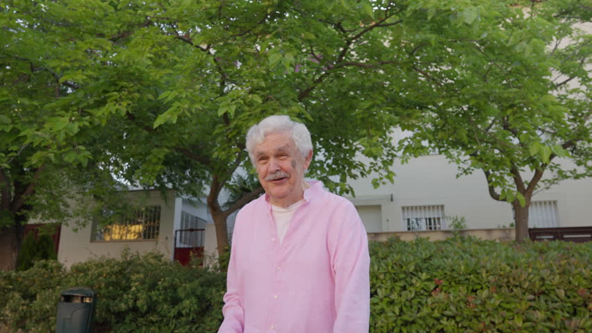 Charming senior man from Madrid, dressed in a light pink shirt, smiles with eyes closed and gestures expressively while sharing a humorous story with an unseen companion in a leafy residential park.