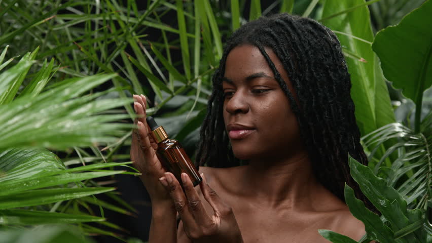 Portrait of beautiful African American woman with long dreadlocks standing in lush jungle greenery, holding face serum bottle and posing for camera