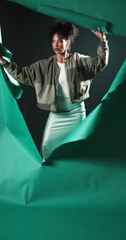 Vertical video: Woman tearing through green paper backdrop, expressing curiosity and exploration. adventure, innovation, breakthrough, creativity, discovery, mystery