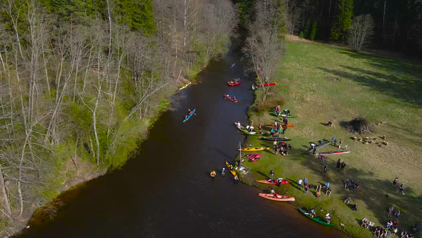 Aerial drone footage flying along kayaks, rafts and SUP paddleboards that are rowing and paddling forward on a narrow dark river during spring sunny day at a kayaking marathon called Võhandu marathon.