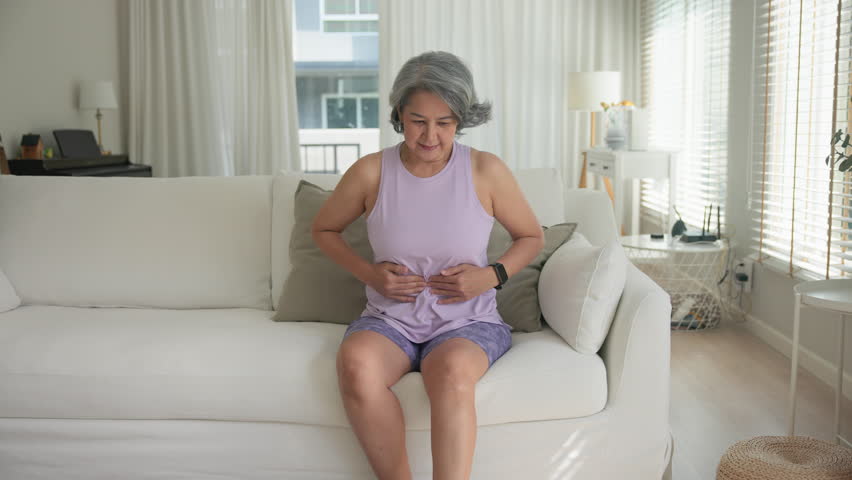 Asia adult middle age woman sit at sofa home feel pain elder gut health care aging system medical issue. Asian female old people Suffer stress cramp abdomen ache cancer illness menses period injury.