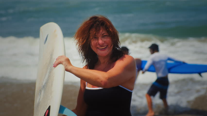 Active mature woman in wetsuit with surfboard at the beach, enjoying a healthy lifestyle. Old senior female doing surfing on Bali. Elderly lady holding surf board. Tropical coast in asia, big wave.