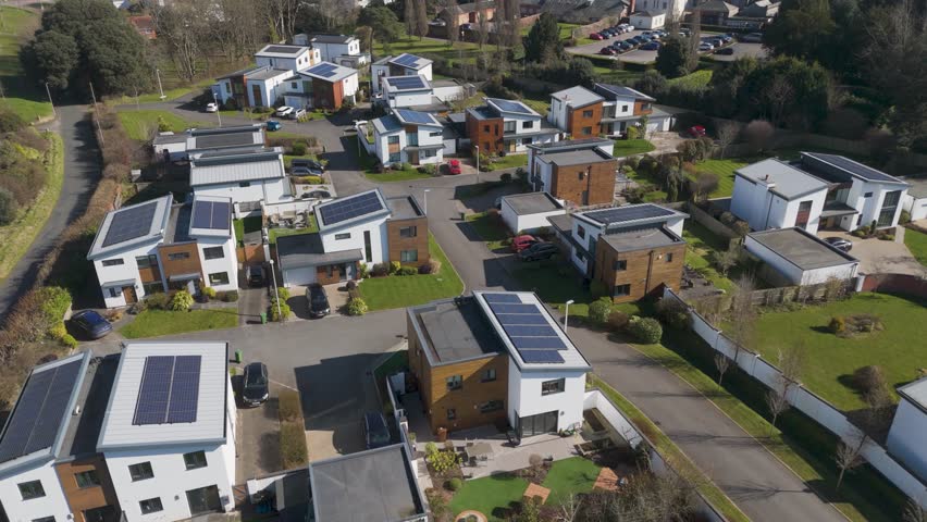 Aerial orbital view of upscale sustainable housing development with angular timber and white facades, rooftop solar panels, geometric landscaping and open green space nearby