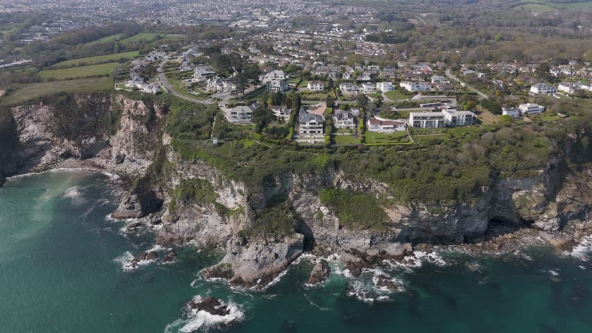 Aerial view of cliff-hugging luxury holiday villas overlooking turquoise Atlantic waves, rugged Cornish coastline and dramatic rock formations, showcasing upscale beachfront living