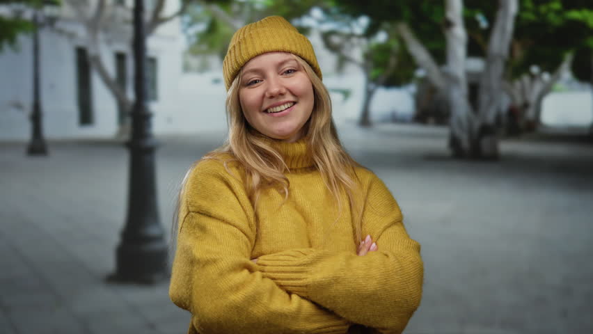 Woman smiling confidently on a city street wearing a yellow sweater and beanie, surrounded by trees and lampposts