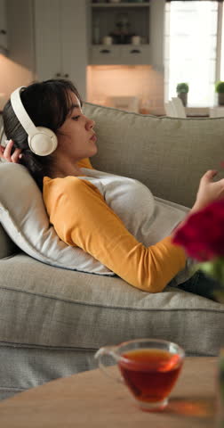 Vertical video: Relaxing on sofa, Asian woman enjoying music with headphones at home. Lifestyle, relaxation, leisure, technology, cozy, unwinding