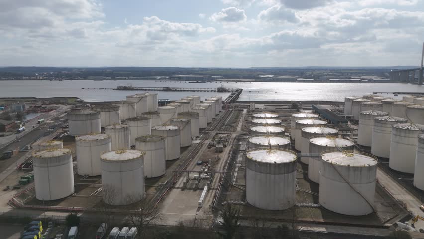 Aerial perspective of expansive riverside fuel storage facility with uniform pale cylindrical tanks stretching into distance under overcast sky, reflecting large-scale energy infrastructure