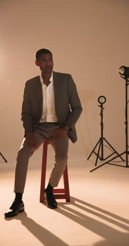 Vertical video: Confident man in stylish suit sitting on stool in professional photography studio. Fashion, confidence, menswear, luxury, handsome, lifestyle