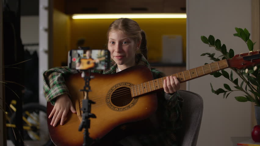 Happy talented Caucasian little girl teen child kid blogger influencer schoolgirl at home recording online video on mobile phone camera tripod playing acoustic guitar singing lesson play music song