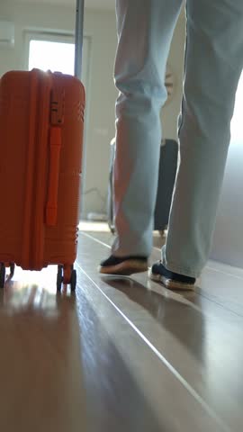 Back view of traveler walking towards door with rolling luggage. Person walks across hallway with orange suitcase. Concept of departure and leaving home for vacation