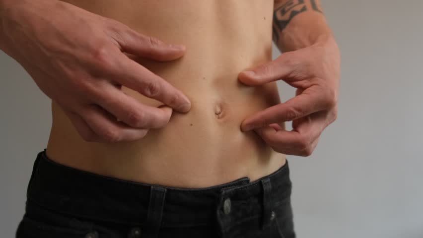 Close-up of fit young man pinching belly skin to show low body fat. Minimal fat layer, lean physique, and healthy abdominal skinfold on gray background.