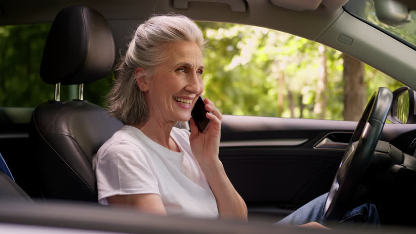 Glad joyful Caucasian elderly mature female woman happy senior lady buyer driver car owner pensioner rental service client using mobile phone calling smartphone talking call inside automobile vehicle