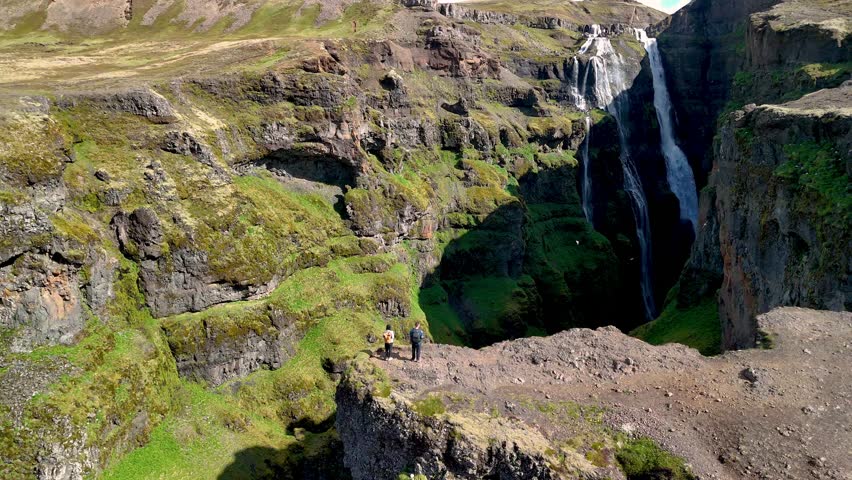 Two hikers stand on a rocky cliff overlooking an impressive waterfall cascading down green cliffs in Iceland. The majestic natural beauty of this remote location, Glymur waterfall canyon Iceland