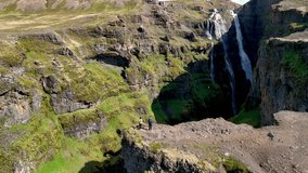 Two hikers stand on a rocky cliff overlooking an impressive waterfall cascading down green cliffs in Iceland. The majestic natural beauty of this remote location, Glymur waterfall canyon Iceland - Powered by Shutterstock - Get 15% off with code: PIKWIZARD15