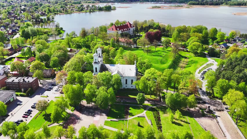 A white church and red-roofed estate rise from a lush riverside park, surrounded by winding paths, city buildings, and shimmering spring foliage in a European town.