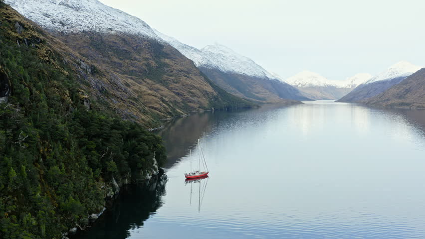 Sailboat travels between narrow fjords in Beagle Channel with high cliffs and smooth water, establishing descend aerial